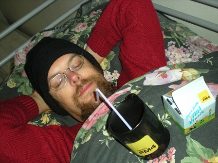 © Bernhard Hilzensauer Mann liegt mit Kappe und geschlossenen Augen auf seinem Bett. Auf seiner Brust eine FM4-Tasse und ein FM4-Kartonradio.